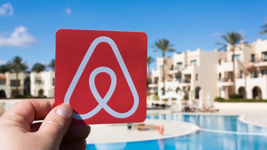 Hand holding an image of the AirBnB logo in front of a swimming pool