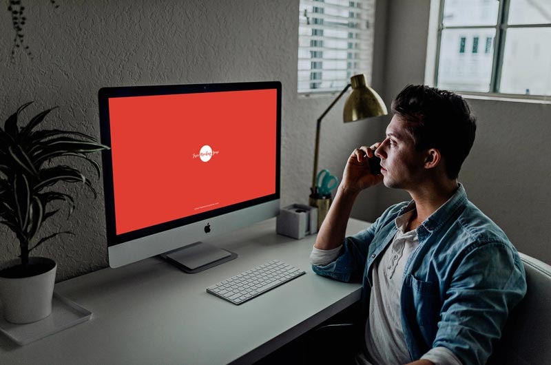 man sitting at a desk, looking at an iMac screen showing a white logo on a red background