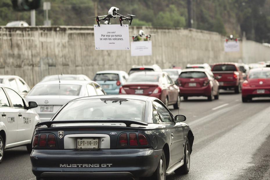 car stopped in traffic, a drone holding a sign in front of the windshield