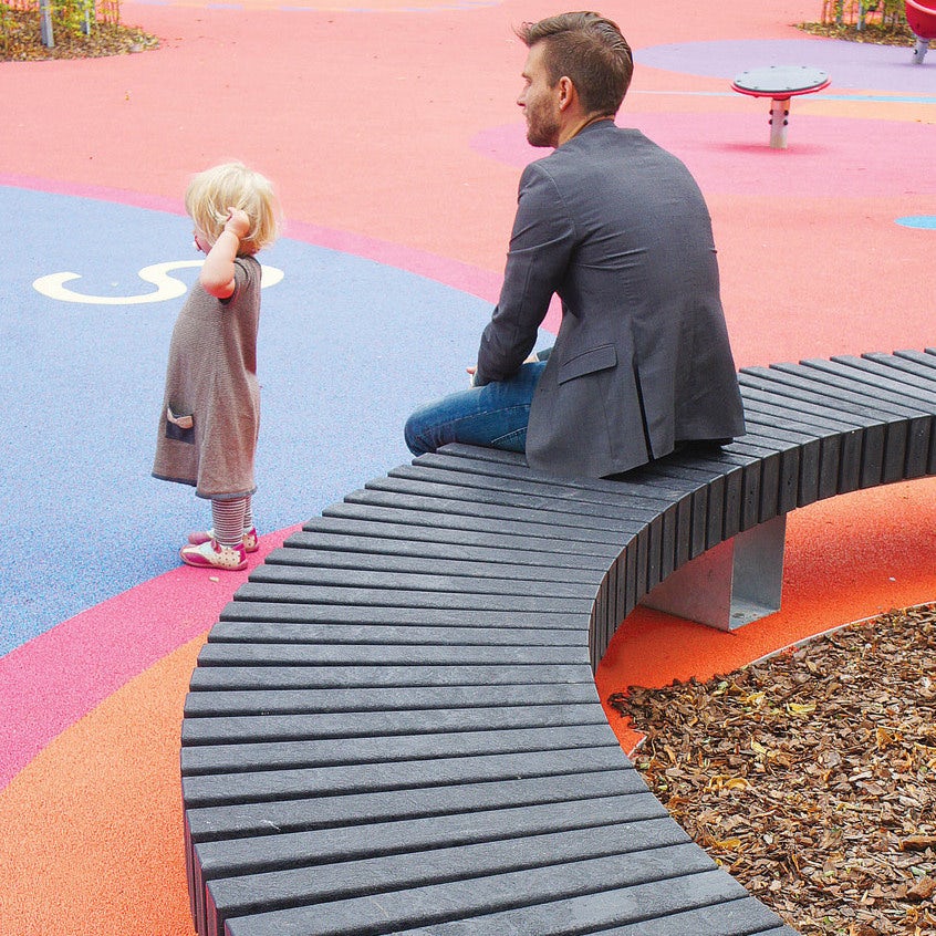 father sitting on a plastic playground bench while watching his child