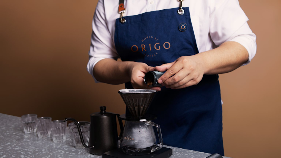 a barista wearing a blue apron pours ground coffee into a filter for brewing