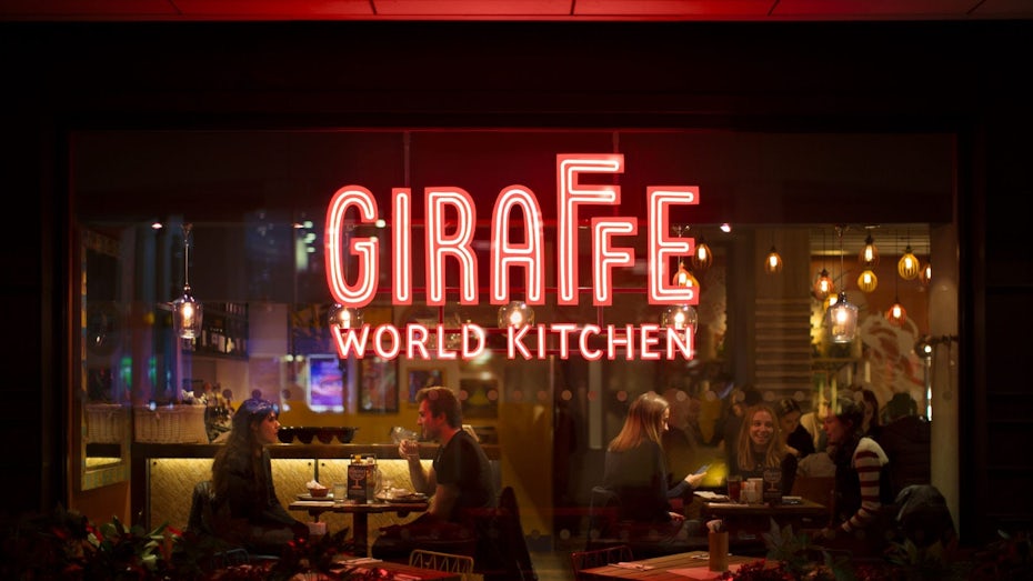 a glass window from the outside looking into a restaurant with a large Giraffe World Kitchen sign in red neon lights