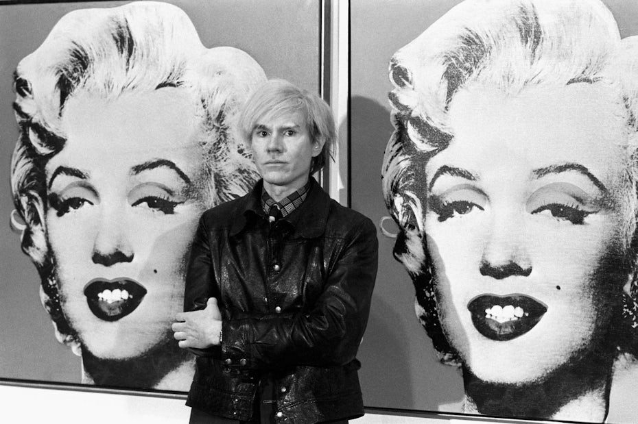 Photograph of Andy Warhol standing in front of his Marilyn Monroe paintings